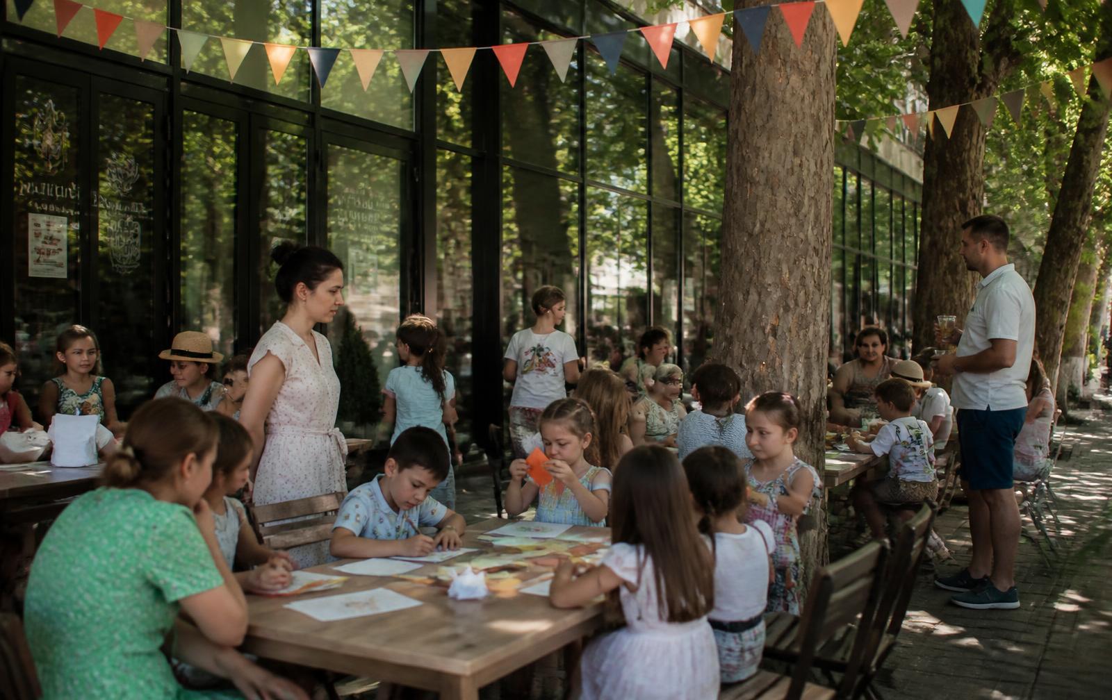 Familii la mese în aer liber, la umbra platanilor, lângă fațada de sticlă a Palatului Național: copii care desenează, fac origami, beau limonadă și mănâncă înghețată, ghirlande de stegulețe de hârtie deasupra