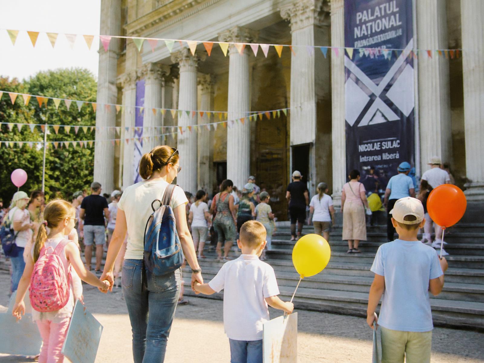 O sală culturală amplă din Chișinău, plină de copii și familii: expoziție de lucrări ale copiilor pe pereți albi, mese de ateliere creative, ghirlande de stegulețe și baloane