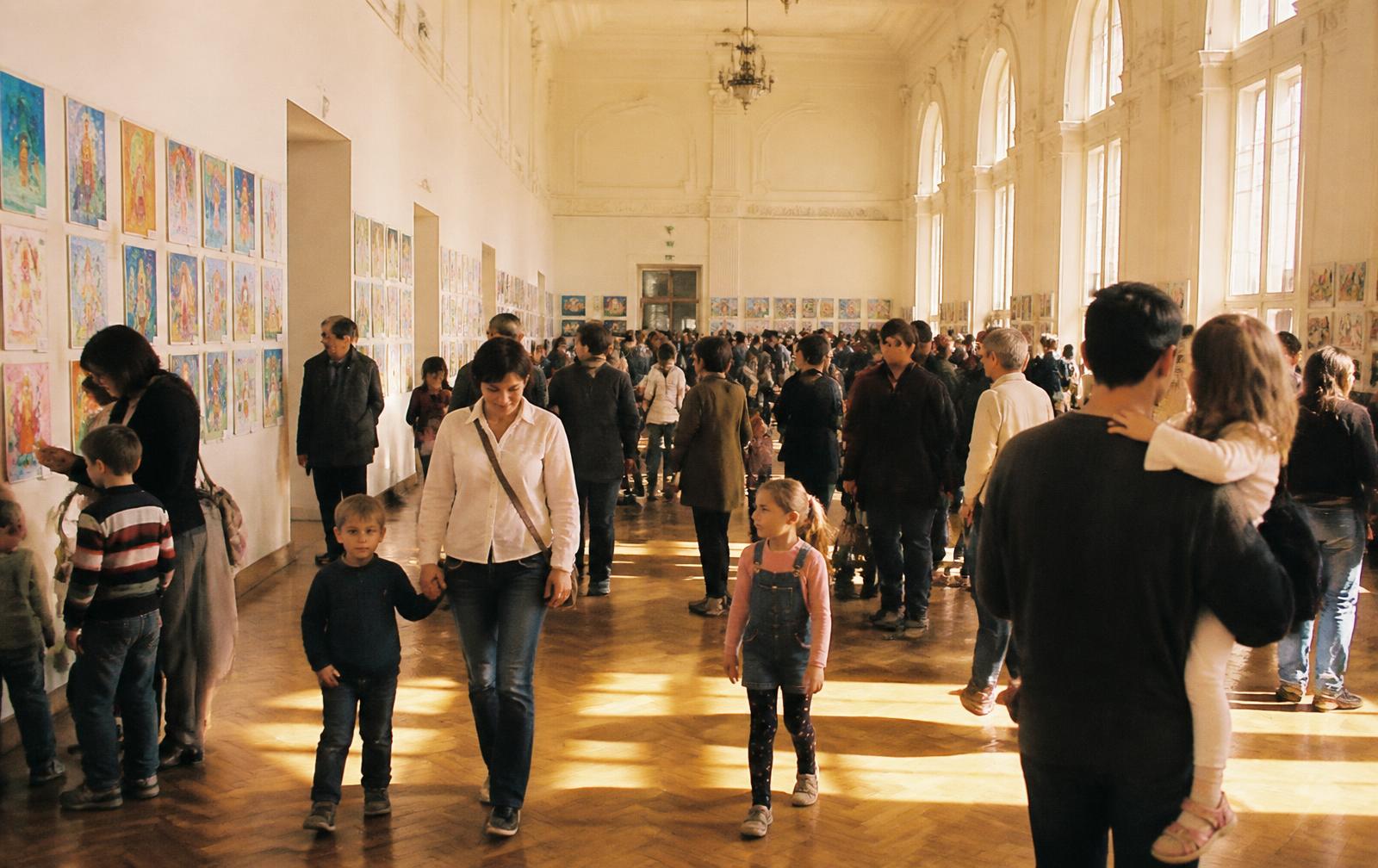 O sală culturală amplă, plină de familii și copii, cu lucrări de copii pe pereți albi