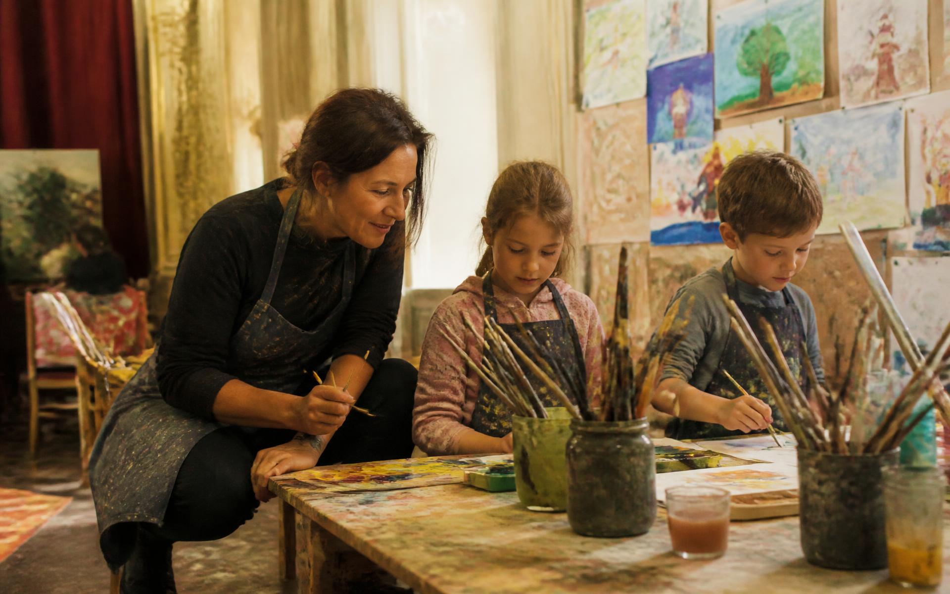 O profesoară de artă lucrează cu doi copii la o masă de atelier plină de vopsea, desene de copii pe perete